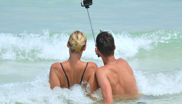 Zwei Personen sitzen am Meerufer in den Wellen und nehmen ein Selfie mit einem Selfiestick auf.