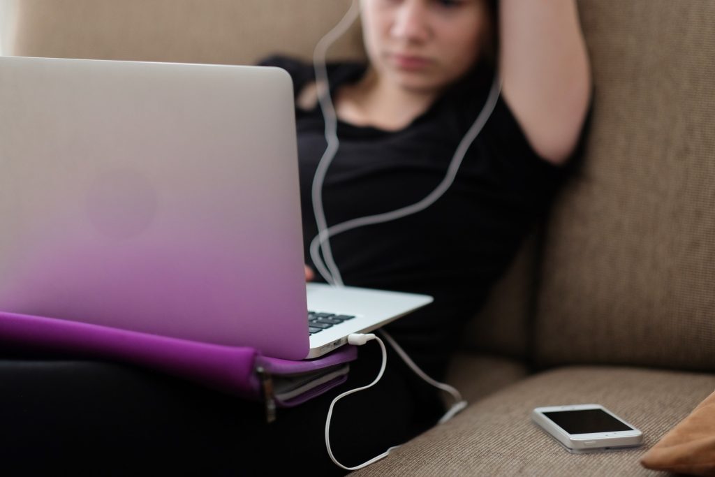 Das Laptop auf dem Schooß im Fokus, verschwommen im Hintergrund ein Mensch mit Kopfhörer und Handy