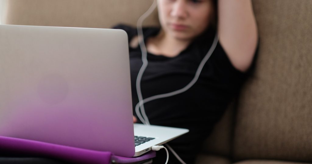 Jeune fille installée sur son canapé regardant l'écran de son ordinateur avec des écouteurs dans les oreilles
