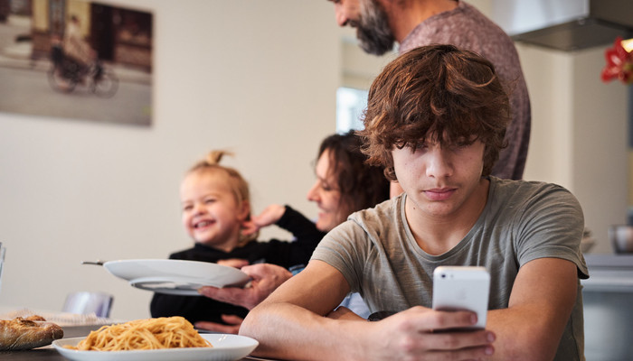 Ein jugendlicher Junge der mit der Familie auf dem Tisch sitzt und am Handy spielt