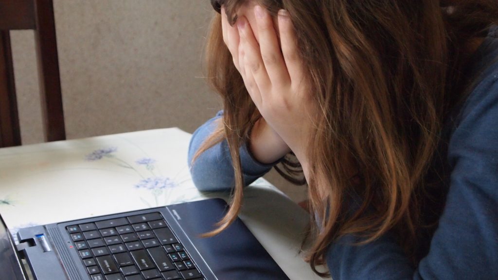 Person mit Langen Haaren, hällt sich, vor dem Computer, mit geneigtem Kopf die Hände vor das Gesicht