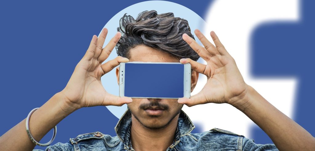 Jeune homme cache ses yeux avec son smartphone, dans l'arrière-plan il y a le logo de Facebook