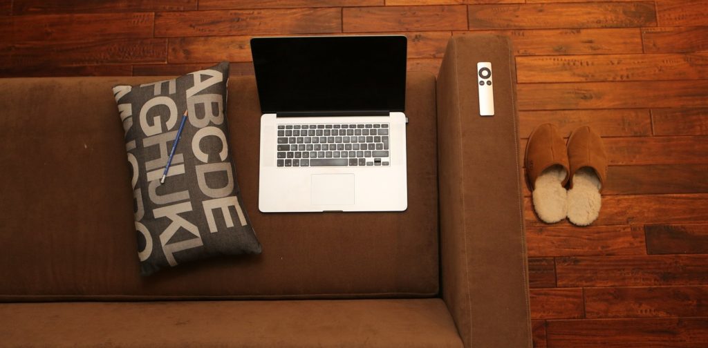 Vue de haut d'un ordinateur portable posé sur un canapé avec à côté un coussin et une télécommande, le tout sur fond marron