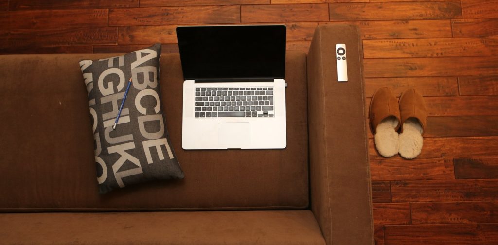 home-office Vue de haut d'un ordinateur portable posé sur un canapé avec à côté un coussin et une télécommande, le tout sur fond marron