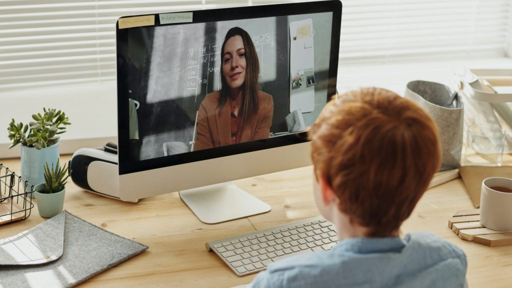 enfant assis devant un bureau en train de regarder un ordinateur avec sa professeure à l'écran