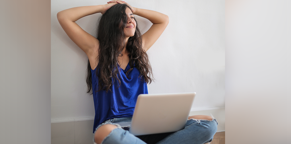 Eine junge Frau sitzt in einem Anzug auf dem Boden gegen der Mauer und schaut lächelnd auf ihren Laptop auf den Knien.