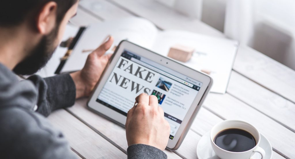 Homme regardant sa tablette avec une page d'un journal ouverte et l'inscription Fake News en grand, un café posé sur la table à côté de lui