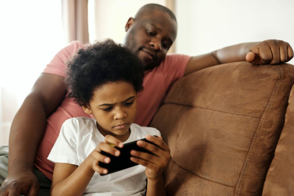 kleiner Junge spielt an seinem Telefon mit seinem Vater, der hinter seine Schulter schaut