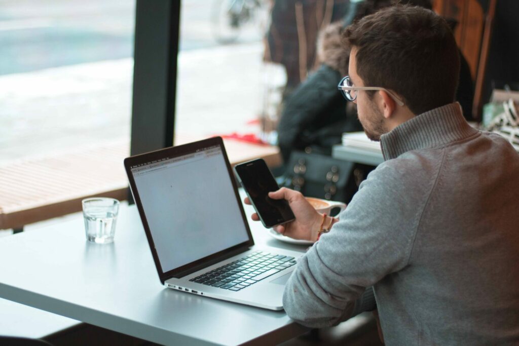 Mann mit Brille sitzt an einem Tisch mit Laptop und schaut auf sein Smartphone