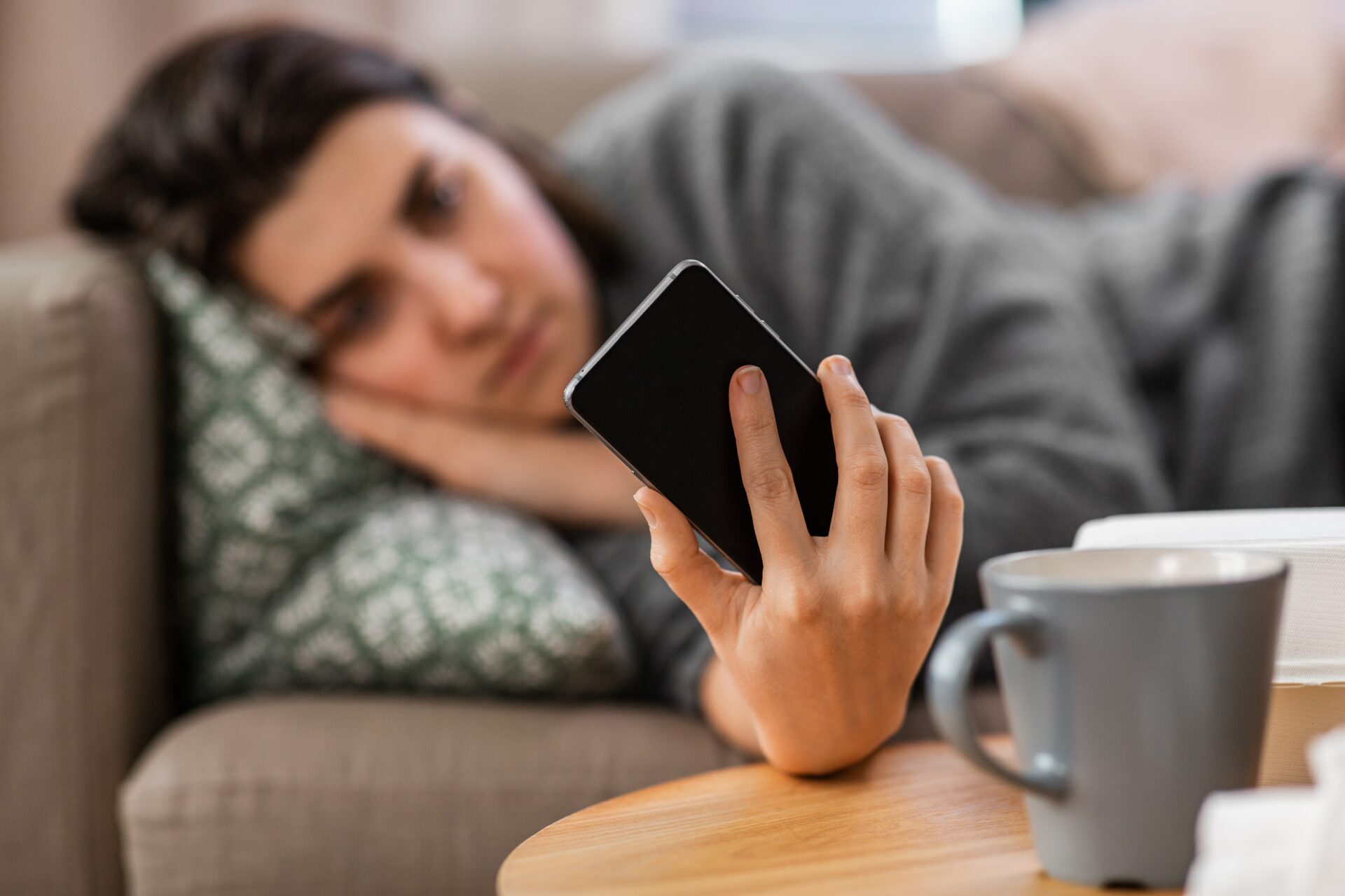 Junge Frau mit Handy in der Hand beim Doom-scrolling auf der Couch.